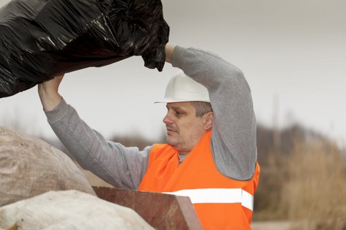 A man wearing a white safety helmet and an orange high-visibility vest is engaged in rubbish removal, lifting a large black plastic bag filled with waste materials. He is outdoors, standing on a practical surface with a background that appears to be an open, rural or semi-urban area with a blurred landscape. The bag is fully expanded, indicating it is filled with waste, and his expression is focused on the task. He is using both hands to lift the bag, which is slightly wrinkled and glossy, suggesting a plastic material. Surrounding him, there are scattered stones, and the environment looks utilitarian, consistent with waste collection or clearance activities. This scene illustrates a professional rubbish removal operation, which could be associated with companies like Office Clearance Kingston upon Thames, providing waste disposal or clearance services across Kingston and nearby areas. The natural lighting emphasizes the outdoor setting, and the in-progress action highlights the practical aspect of rubbish handling and transportation.