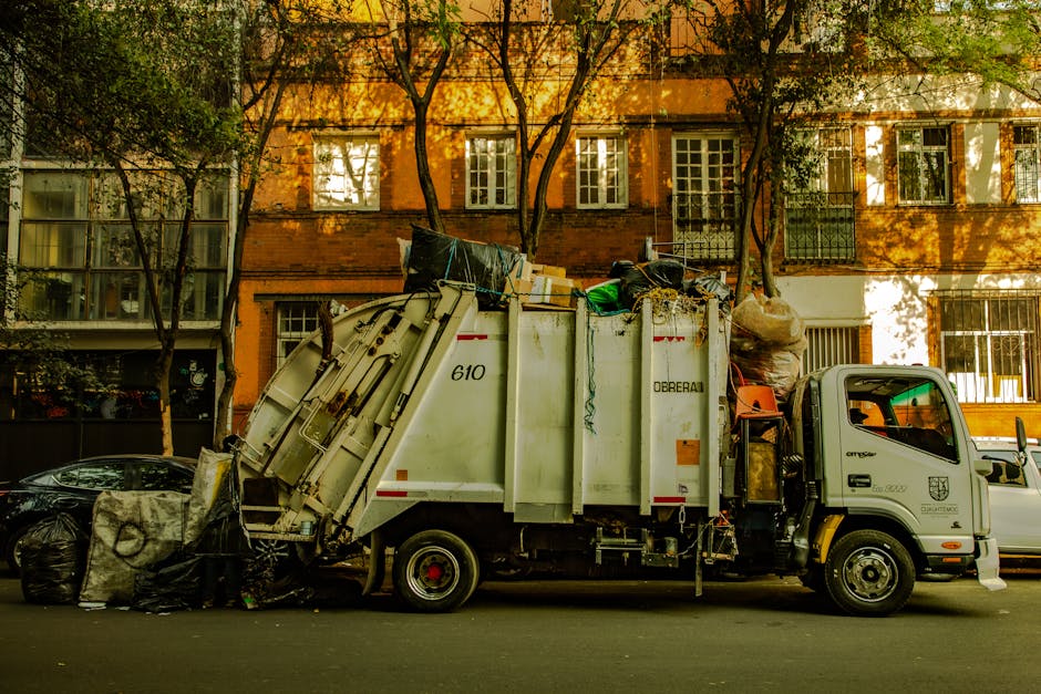 A row of six black plastic rubbish bags, filled and tied, are stacked against the exterior wall of a green wooden building on a concrete sidewalk, near a set of wooden stairs leading up to a white doo