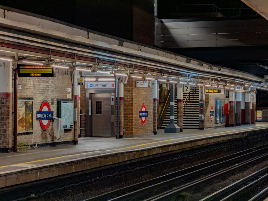 At an outdoor train station platform, a metal recycling and waste disposal unit with four separate colour-coded compartments is prominently positioned in the foreground. The unit features clearly labe