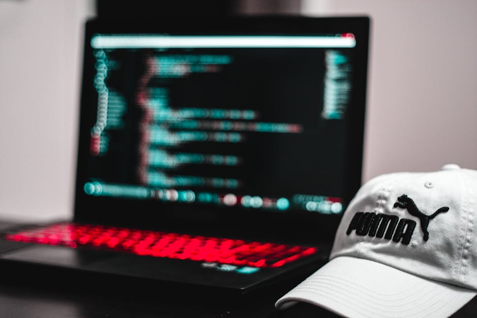 A close-up view of a modern laptop with a black body and a red-backlit keyboard, positioned on a dark surface in an indoor setting. The laptop screen displays blurred lines of code or text in shades o